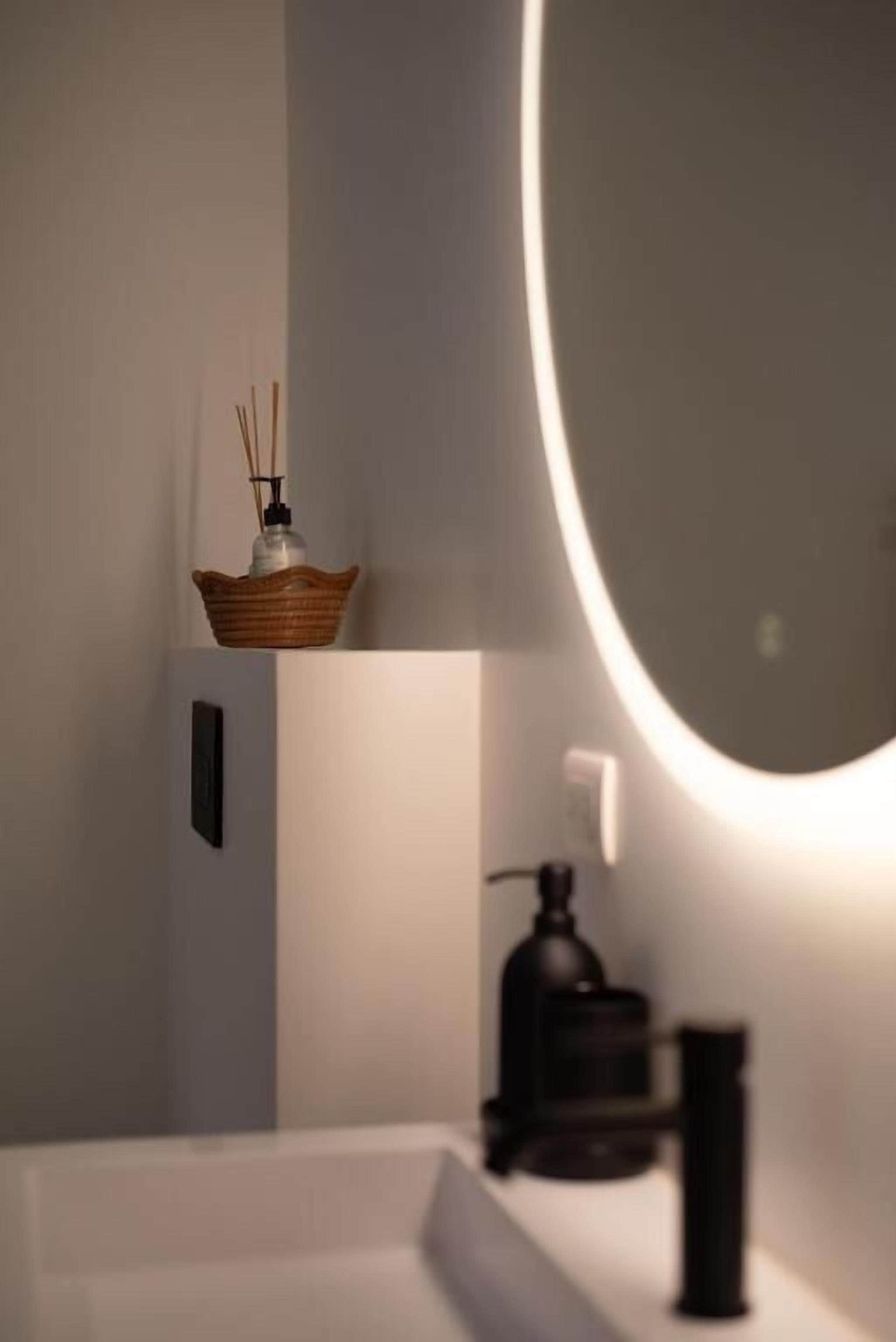 Ein modernes Badezimmer mit einem runden Spiegel, einem weißen Waschbecken, einem schwarzen Wasserhahn und einem dekorativen Korb mit einem Reed-Diffusor.