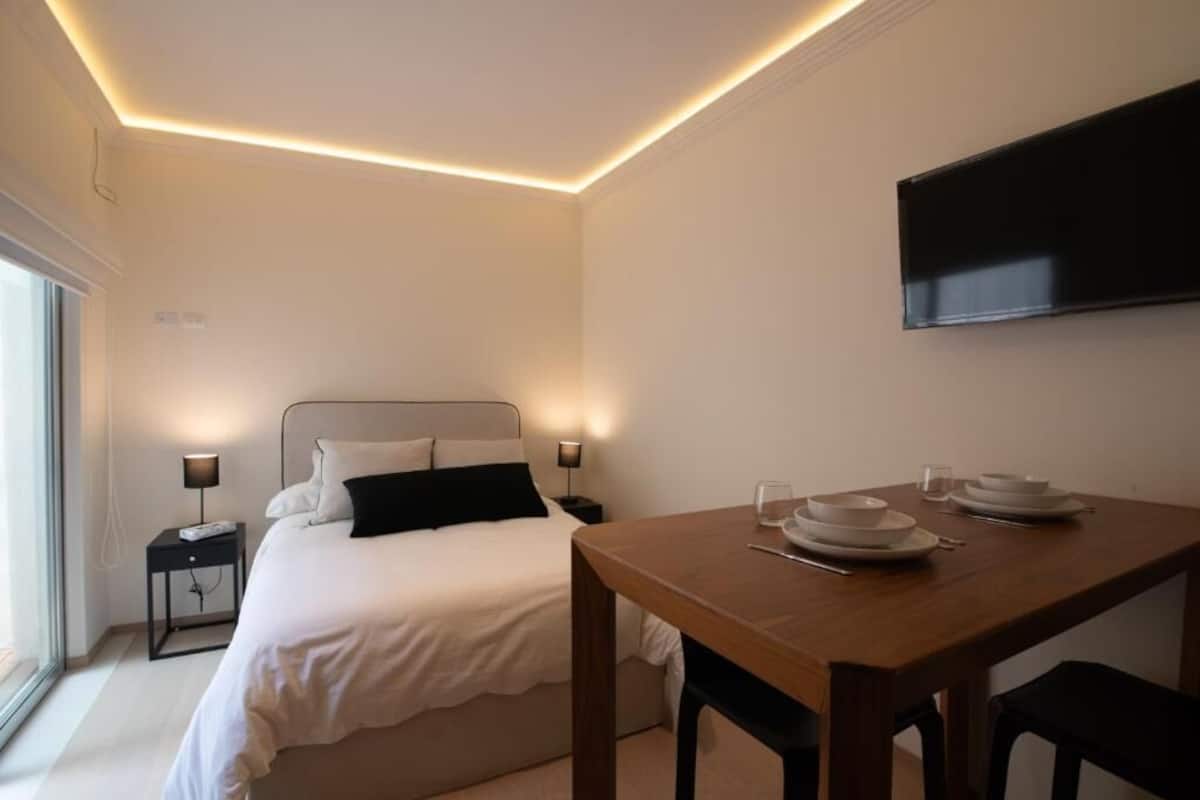 A bedroom with a bed, bedside tables, a TV mounted on the wall, and a dining table with chairs.