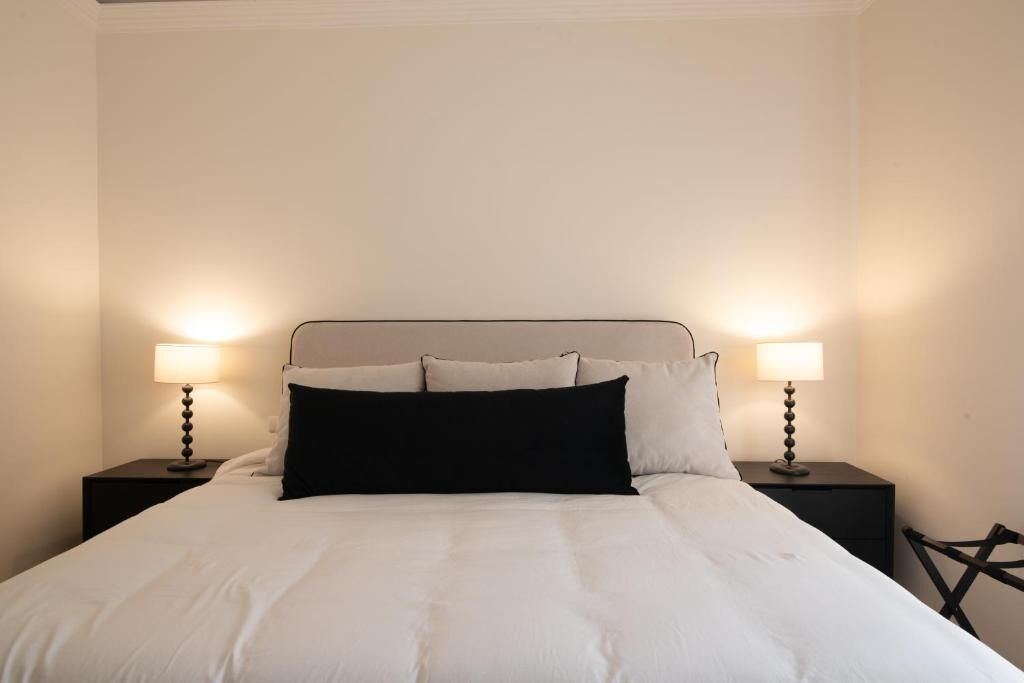 A bed with white bedding and black pillows, flanked by two bedside tables with lamps, in a room with plain walls.