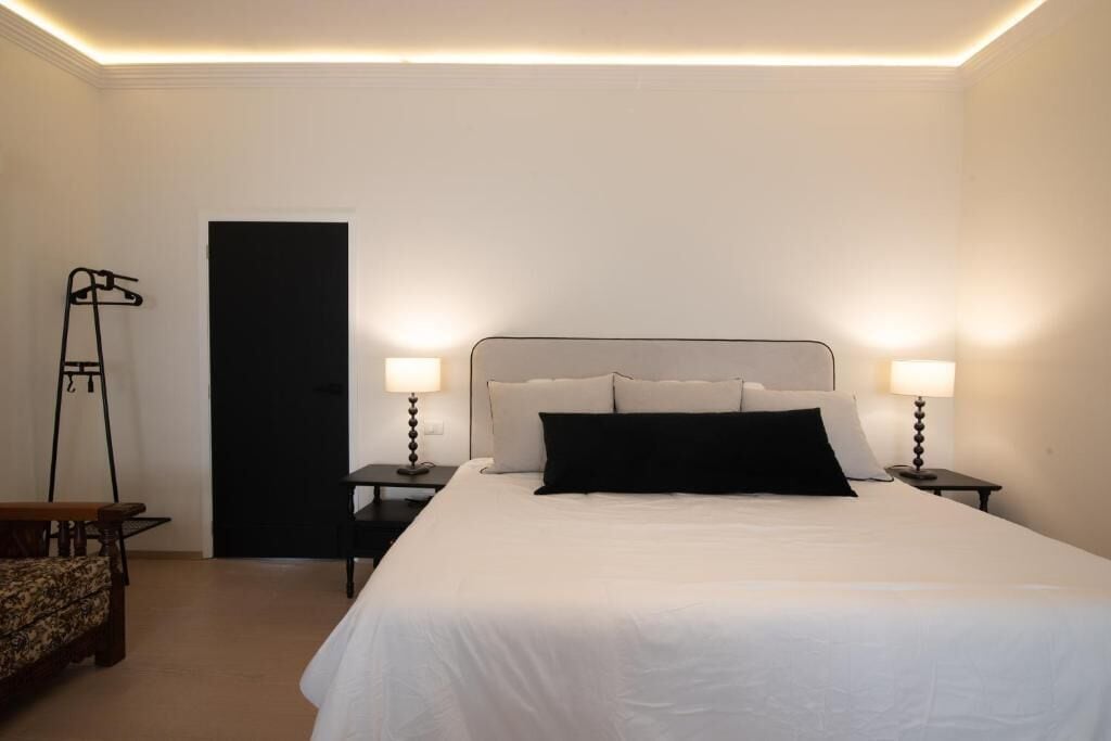 Una habitación de hotel con cama, dos mesitas de noche, un sillón y una puerta.