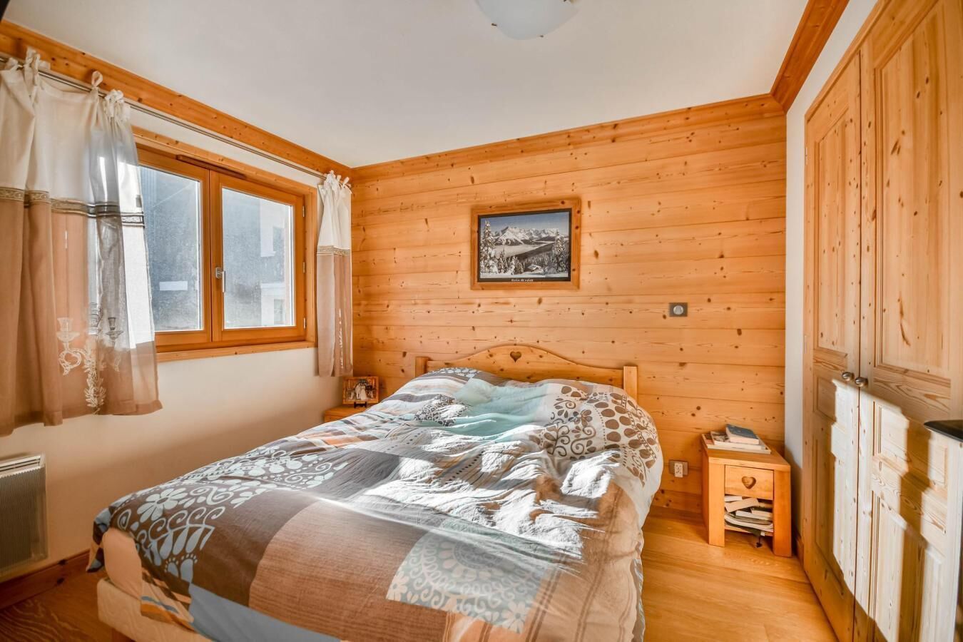 Un dormitorio con paneles de madera, una cama, una mesita de noche, un radiador y una ventana con cortinas.