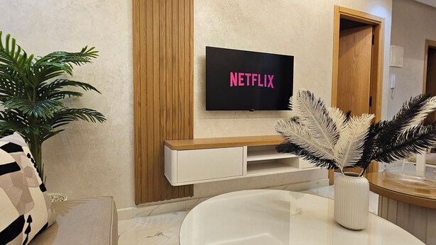 Un salon moderne avec un téléviseur affichant le logo « NETFLIX », un canapé blanc, une grande plante en pot et un vase décoratif orné de plumes.