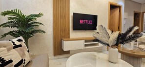 Un salon moderne avec un téléviseur affichant le logo « NETFLIX », un canapé blanc, une grande plante en pot et un vase décoratif orné de plumes.