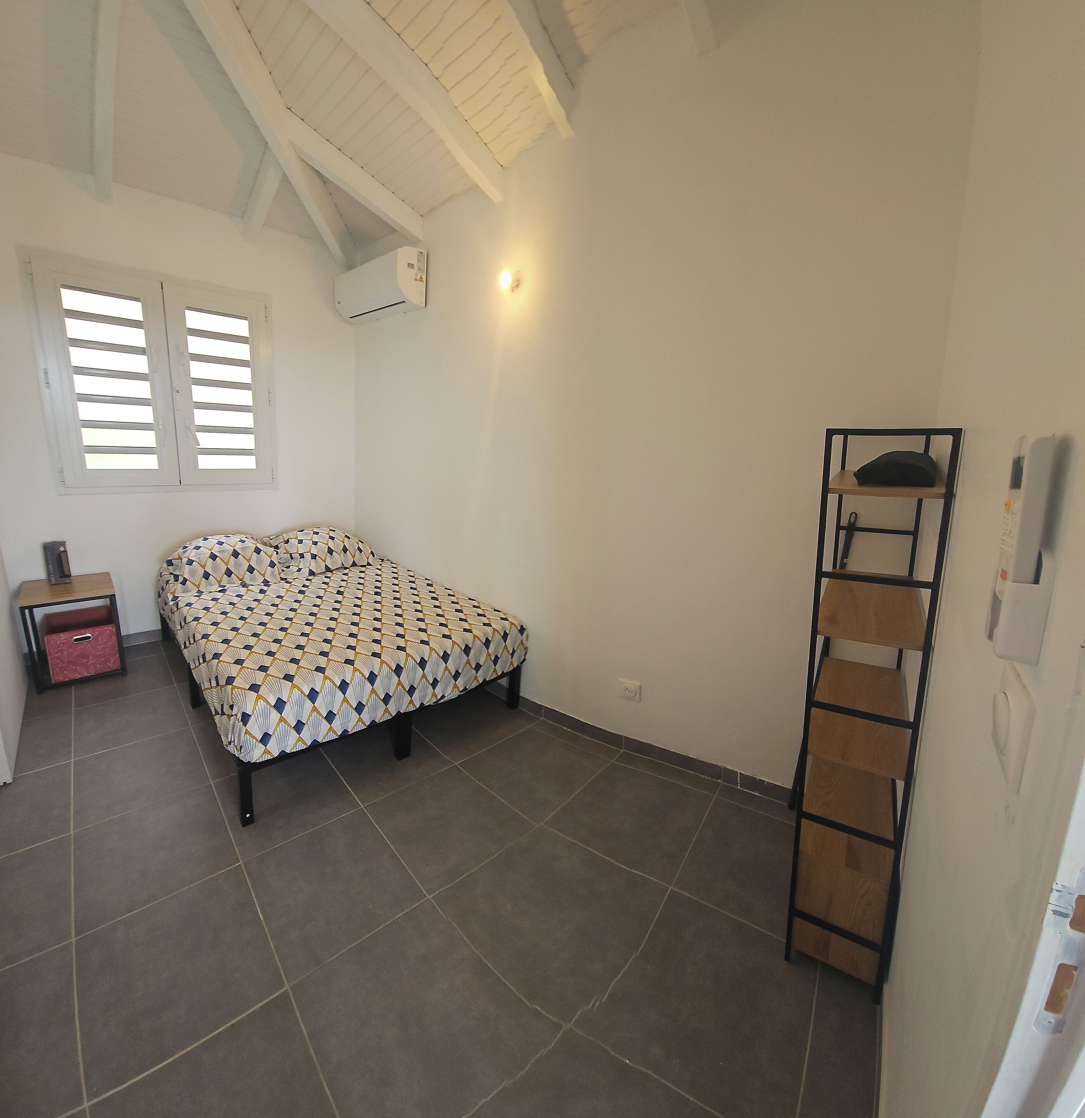 A bedroom with a bed, a nightstand, a red storage box, a wall-mounted shelf, and a ceiling fan.