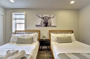 Habitación de hotel con dos camas, una ventana con persianas, un cuadro de un toro colgado en la pared y una mesita de noche con una planta.