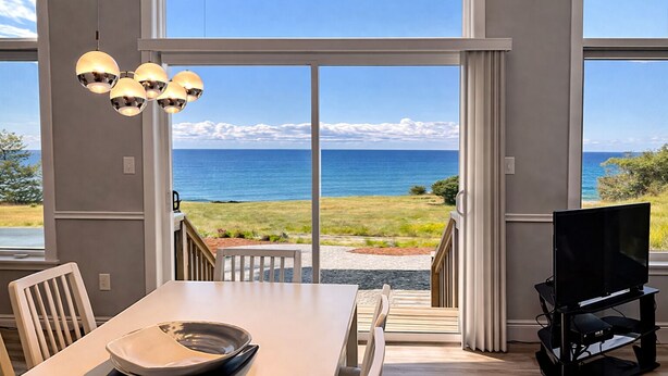 A room with a dining table, chairs, a TV, and a view of the ocean through large sliding doors.