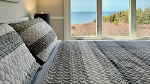 A bedroom with a large bed, a painting on the wall, and a view of the ocean through a sliding door.