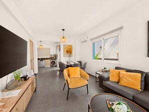 Eine moderne Wohnung mit Wohnzimmer, Essbereich und Küche. Es gibt einen großen Flachbildfernseher, eine Couch mit orangefarbenen Kissen und einen Esstisch mit Stühlen.