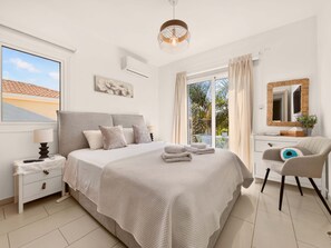 A bedroom with a bed, bedside table, lamp, and a view of the outdoors. - Konnos Haven Villa, Cape Greco Escape (Protaras)
