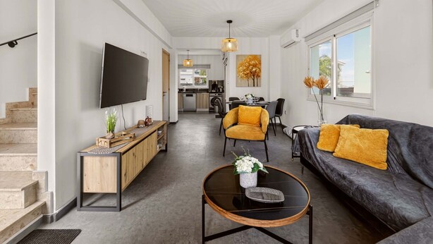 A modern living room with a sofa, coffee table, and TV. There is a staircase on the left and a kitchen in the background. - Konnos Haven Villa, Cape Greco Escape (Protaras)