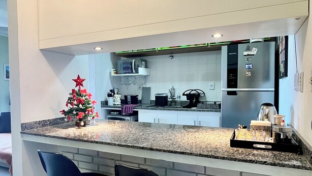 A modern kitchen with a granite countertop, two black bar stools, a stainless steel refrigerator, and a small Christmas tree. - Home office with pool and barbecue in Capão Novo - RS (Capão Novo)