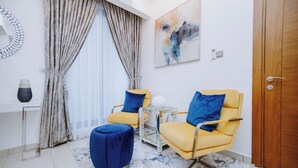 Une pièce moderne avec deux chaises jaunes, un pouf bleu, une table en verre et un tableau accroché au mur.