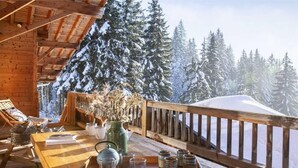 Une terrasse en bois avec une rambarde en bois, une table sur laquelle se trouve un plateau avec des verres et un vase, et une vue sur des arbres couverts de neige.