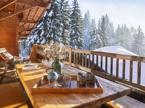 Eine Holzterrasse mit Holzgeländer, einem Tisch mit einer Ablage für Gläser und einer Vase, und Blick auf schneebedeckte Bäume.