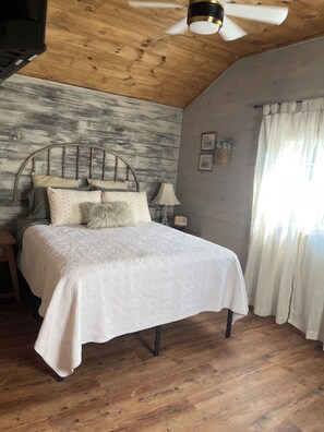Ein Schlafzimmer mit Holzbalkendecke, einem Bett mit weißer Bettdecke, einem hölzernen Kopfende und einem Fenster mit Vorhängen.