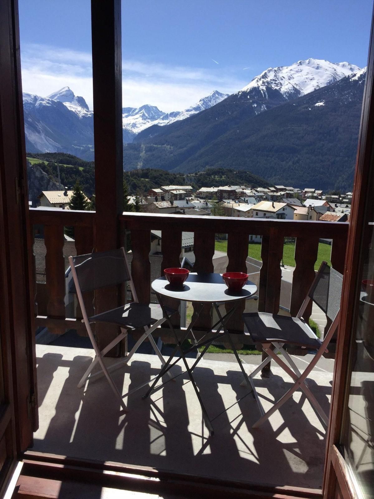 A balcony with a table and chairs, overlooking a mountainous landscape with snow-capped peaks.