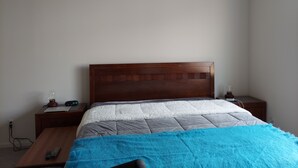 Een slaapkamer met een houten hoofdbord, een bed met een grijs-witte quilt, een blauwe deken, een nachtkastje met een lamp en een afstandsbediening op het nachtkastje.