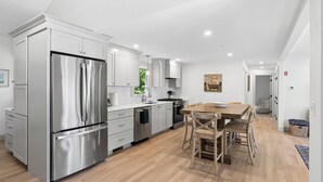 A modern kitchen with stainless steel appliances, white cabinetry, and a wooden dining table with chairs. - Luxe 5 Bed/ 3 Bath with Game Room & AC (Yarmouth)