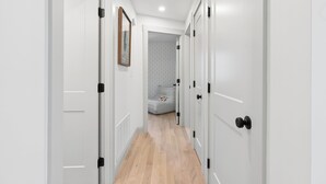 A narrow hallway with white doors, a framed picture on the left wall, and a bed in the background. - Luxe 5 Bed/ 3 Bath with Game Room & AC (Yarmouth)