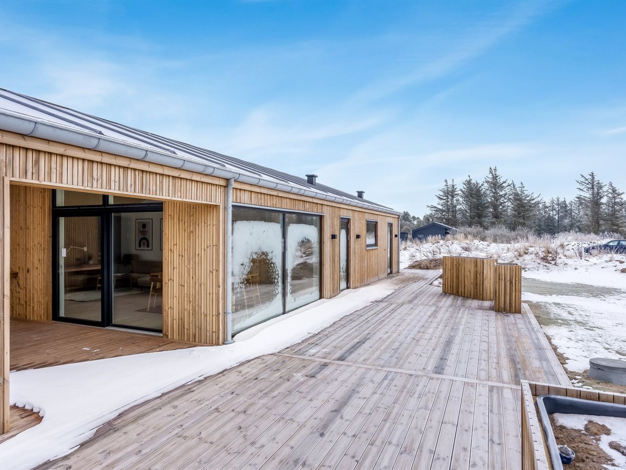 En moderne træbygning med store glasruder, en træterrasse og et sneklædt landskab.