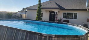 A swimming pool with a wooden deck, a basketball hoop, and a house with a dark roof and a door with a sign. - Villa Privada San Carlos (Ensenada)