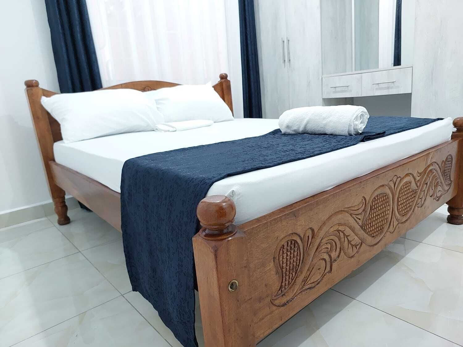 A wooden bed with white bedding and a blue throw blanket, a wooden nightstand, and a white wardrobe with a mirror.
