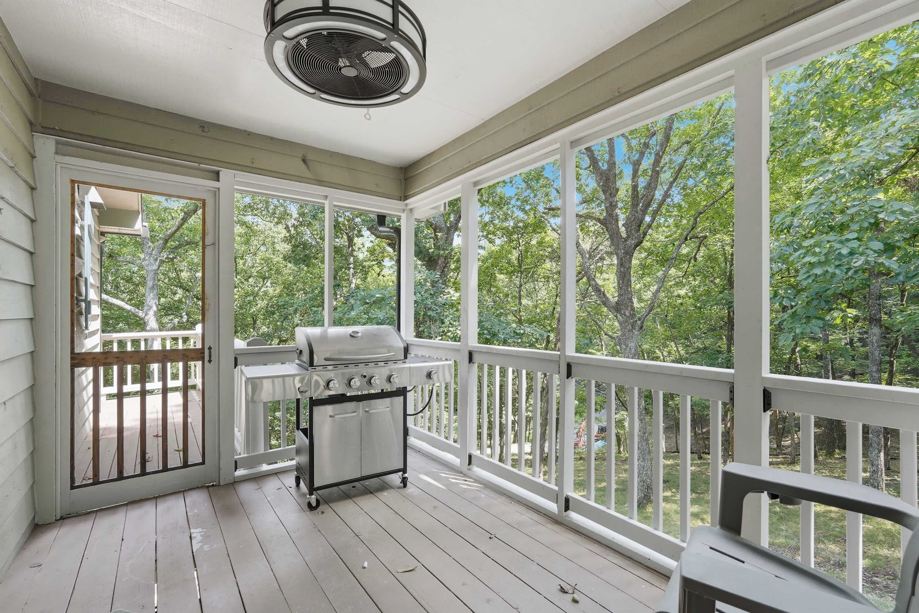 A covered porch with a grill and a fan, overlooking a wooded area.