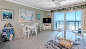 Una sala de estar con televisor, mesa de comedor, sillas, una tabla de surf y un balcón con vistas a la playa.