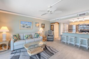 A living room with a sofa, armchairs, a glass coffee table, and a kitchen area with a bar and stools. - Gateway Villas 595 (Fort Myers Beach)