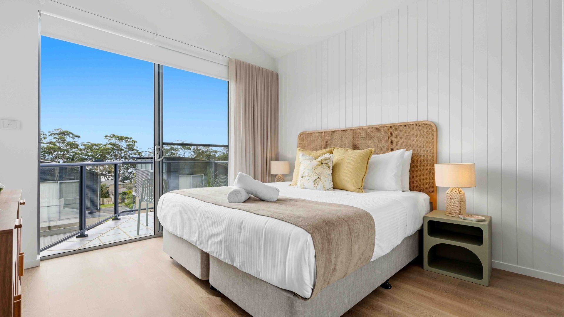 A modern bedroom with a large bed, a nightstand, a lamp, and a view of the outdoors.