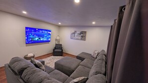A living room with a large flat-screen TV, a gray sectional sofa, a floor lamp, and a painting on the wall.