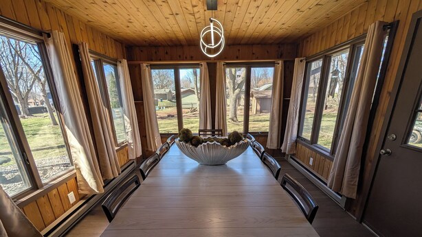 A long wooden dining table with a central bowl, surrounded by chairs, in a room with large windows and wooden walls.