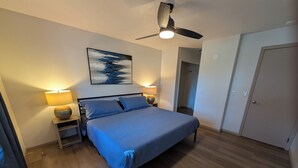 A bedroom with a bed, two bedside lamps, a ceiling fan, and a painting on the wall.