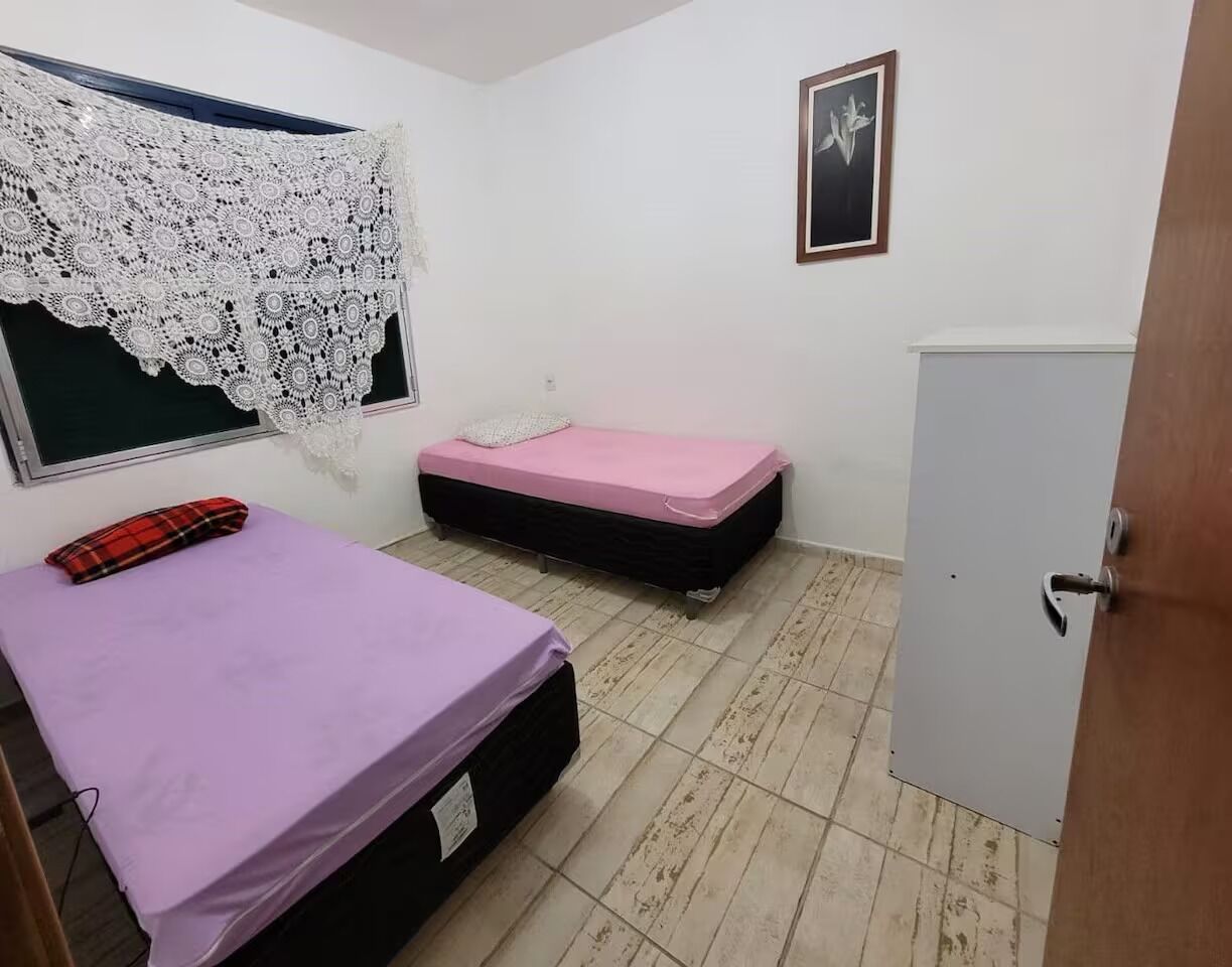 Een kleine kamer met twee bedden, een raam met een wit kantgordijn, een lijst met een afbeelding aan de muur en een deur.