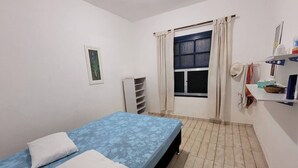 A small, clean room with a single bed, a window with curtains, a radiator, and a shelf with personal items. - Casa em Ilha Comprida (Ilha Comprida)