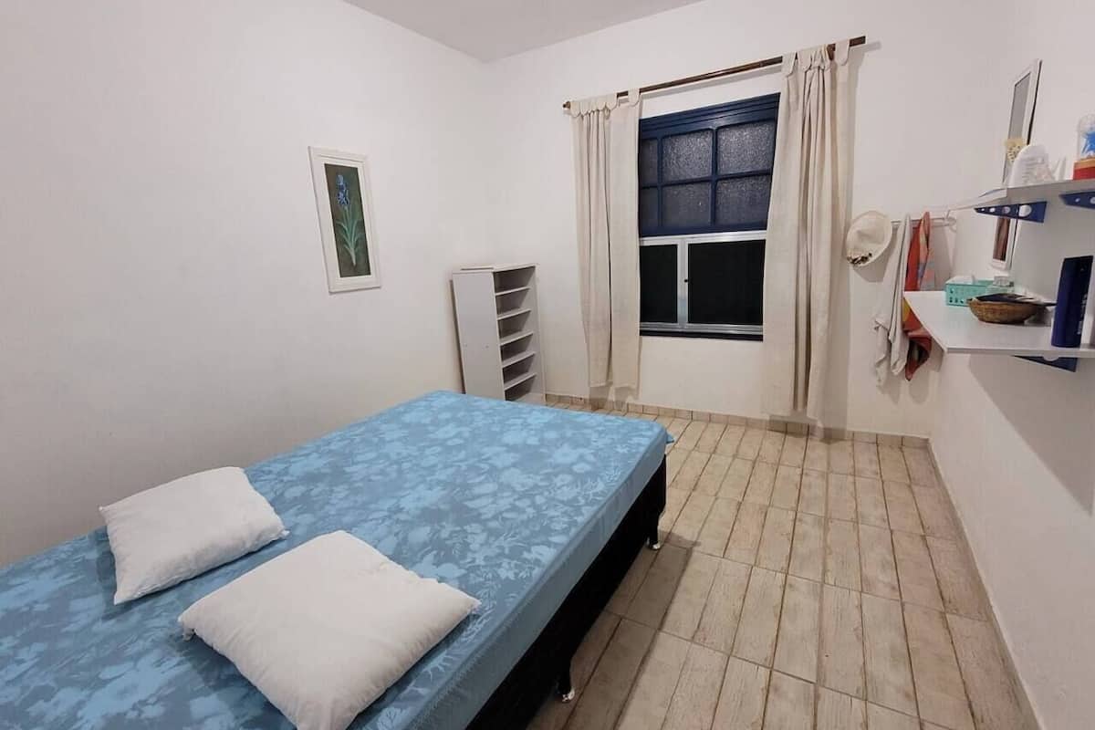 A small, clean room with a single bed, a window with curtains, a radiator, and a shelf with personal items.
