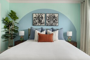 Un lit avec une literie blanche, deux lampes fixées au mur, une plante en pot et deux reproductions botaniques encadrées accrochées au mur.