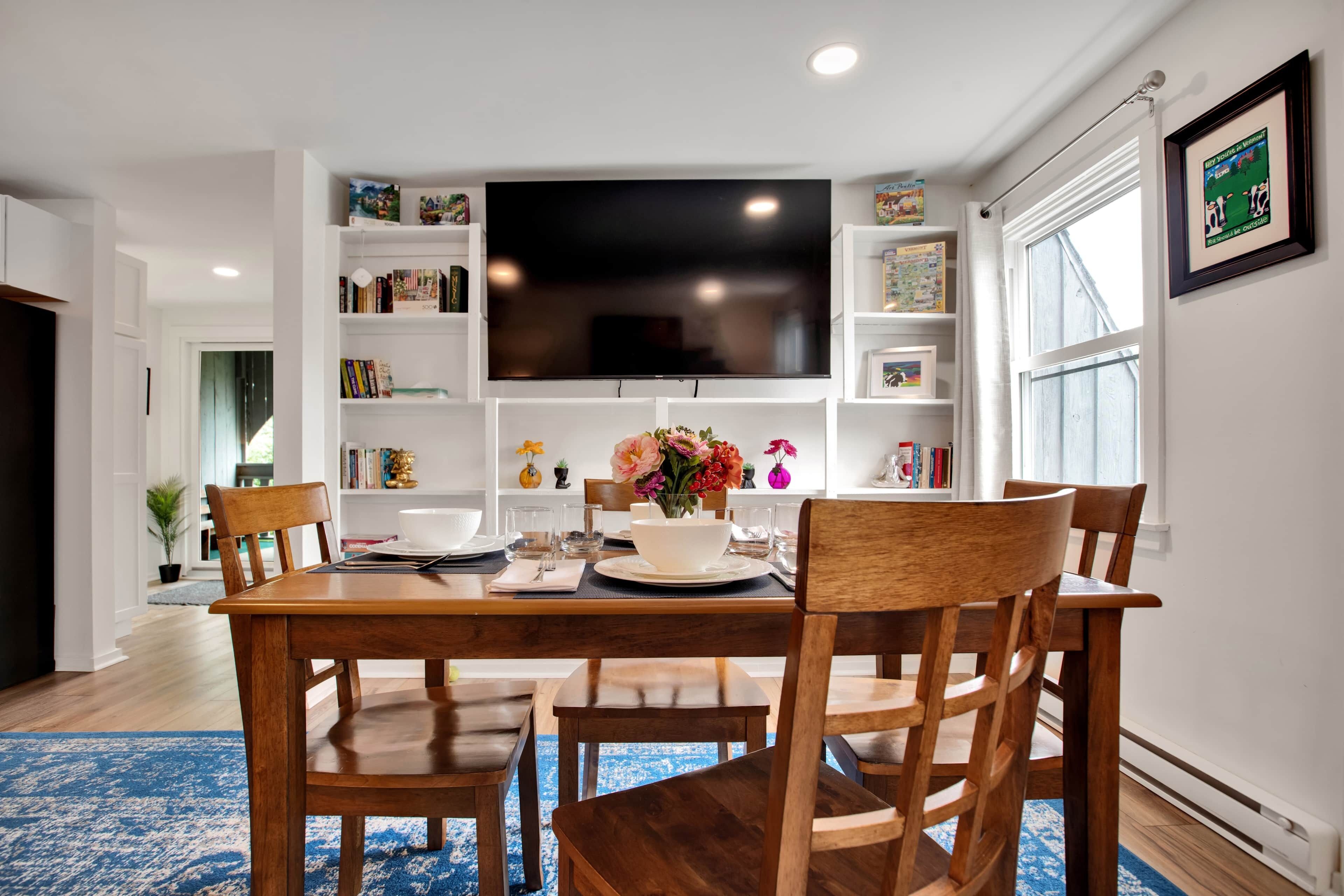 A dining area with a wooden table, chairs, a flat-screen TV mounted above, and bookshelves with various items.
