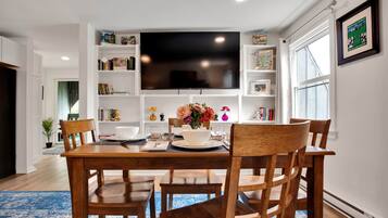 A dining area with a wooden table, chairs, a flat-screen TV mounted above, and bookshelves with various items.