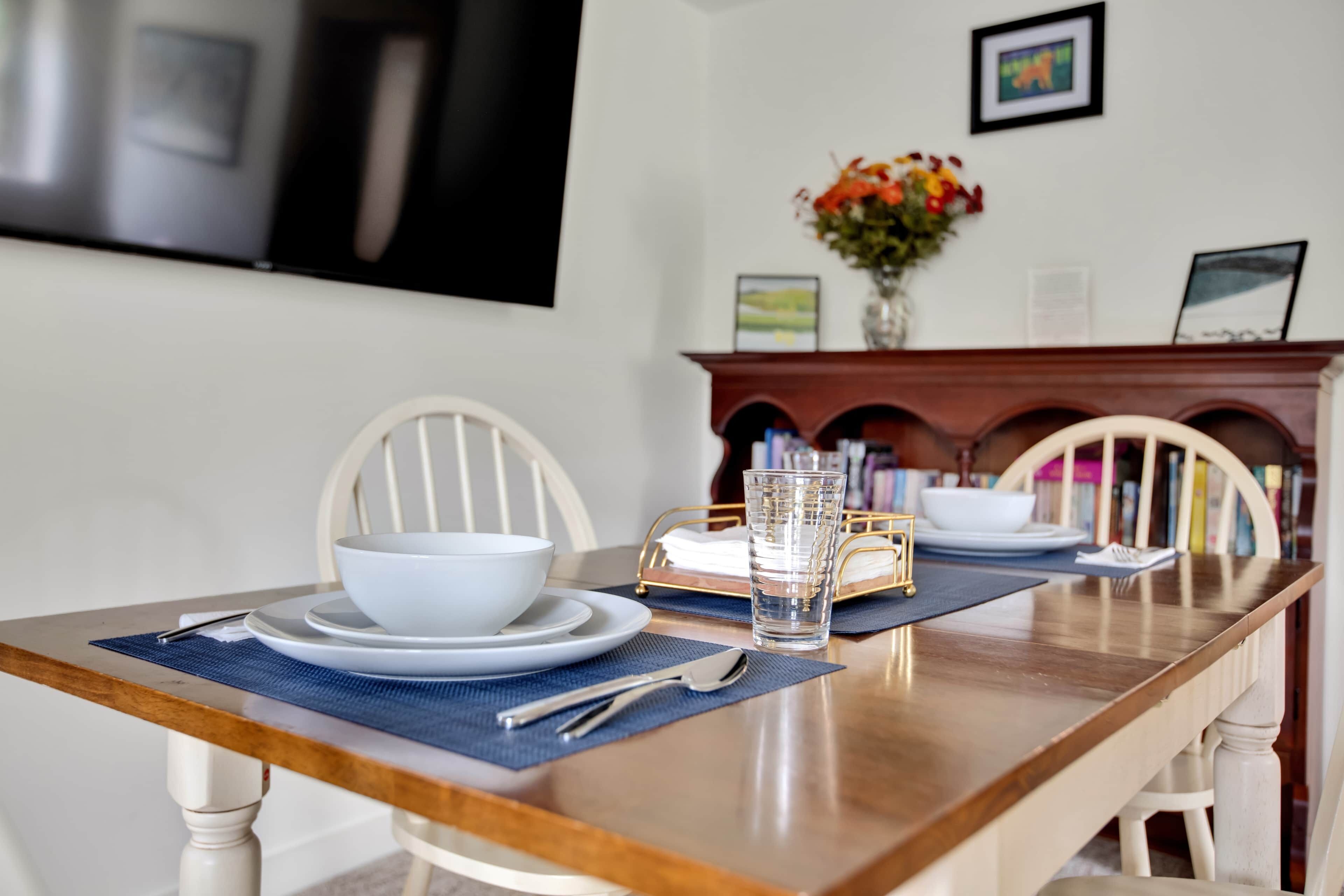 A dining table set with white plates, bowls, and a glass. There are chairs with a curved backrest, a vase with flowers, and a fireplace with a mantel.