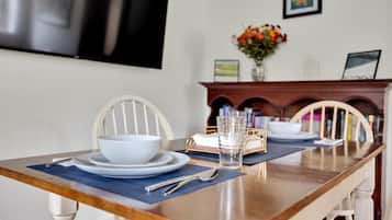 A dining table set with white plates, bowls, and a glass. There are chairs with a curved backrest, a vase with flowers, and a fireplace with a mantel.