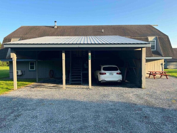 A covered parking area with a car parked inside, a picnic table, and a building with a sloped roof.