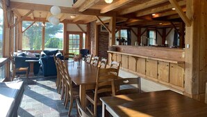 Un comedor espacioso con una larga mesa de madera y sillas, una zona de bar con armarios de madera y ventanas amplias que dejan entrar luz natural.
