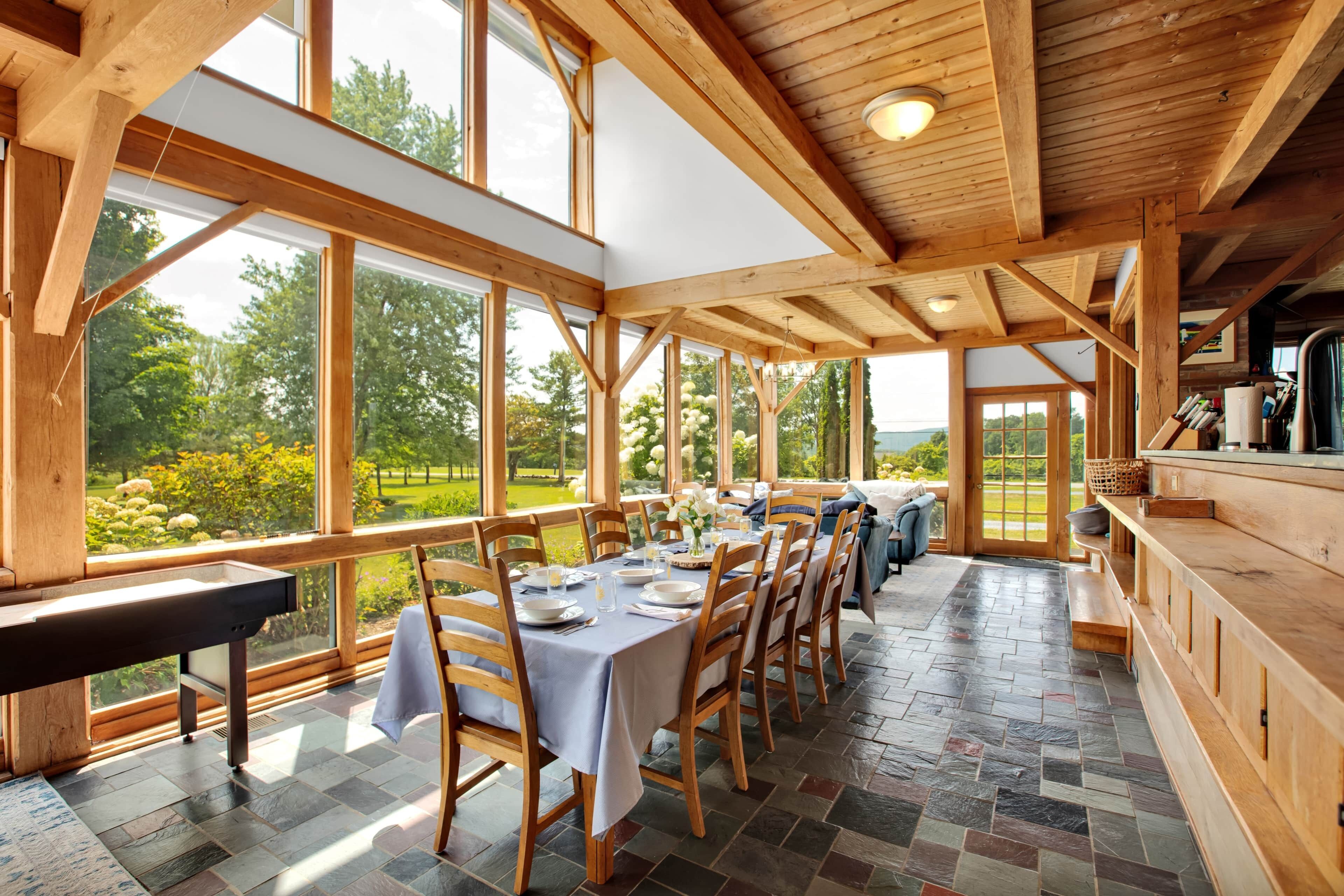 Une salle à manger spacieuse avec une longue table dressée pour six personnes, de grandes fenêtres offrant une vue sur l’extérieur et une structure de plafond en bois.