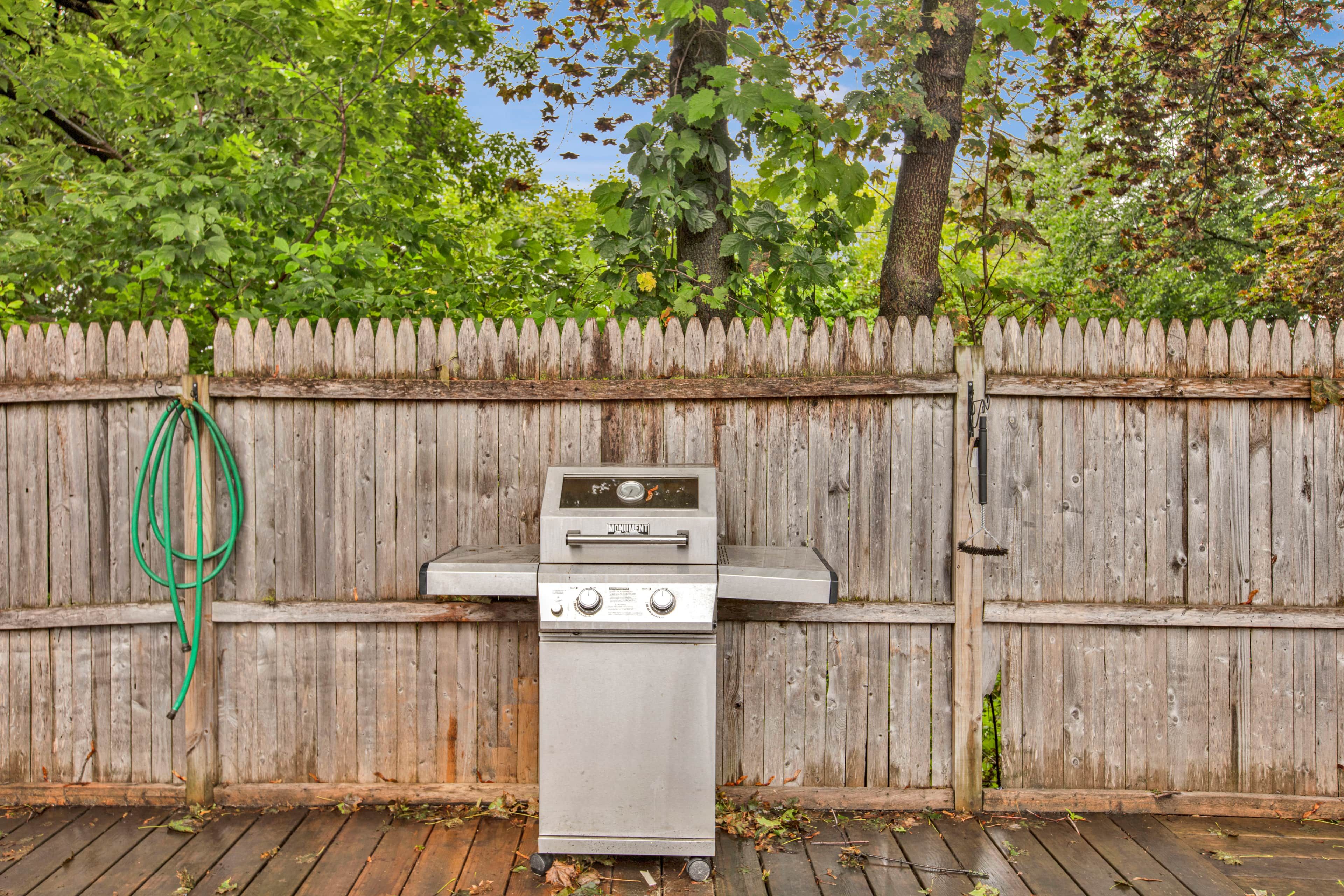 A stainless steel outdoor grill with a control panel, a green garden hose, and a wooden fence in the background.