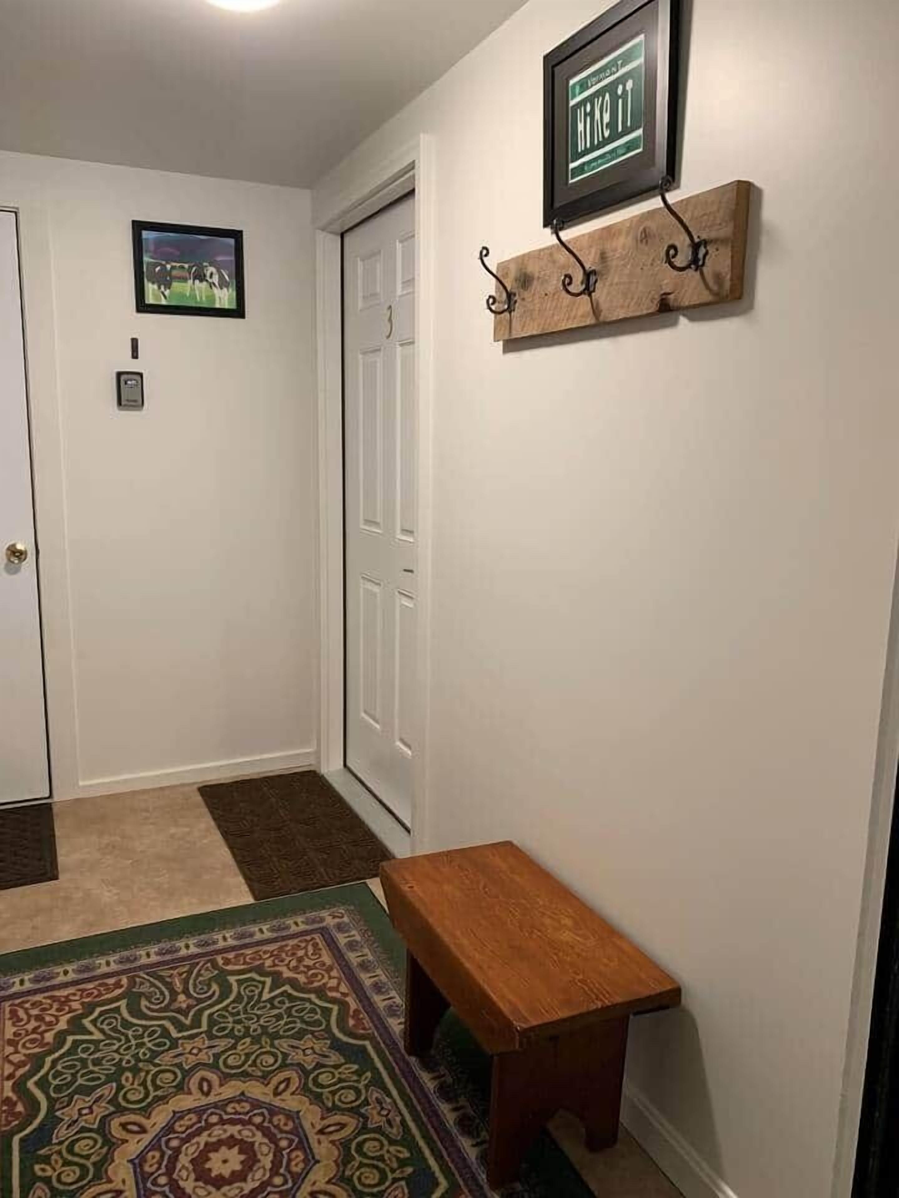 Un pasillo con un banco de madera, un tapete estampado, una puerta con número y un cartel en la pared.