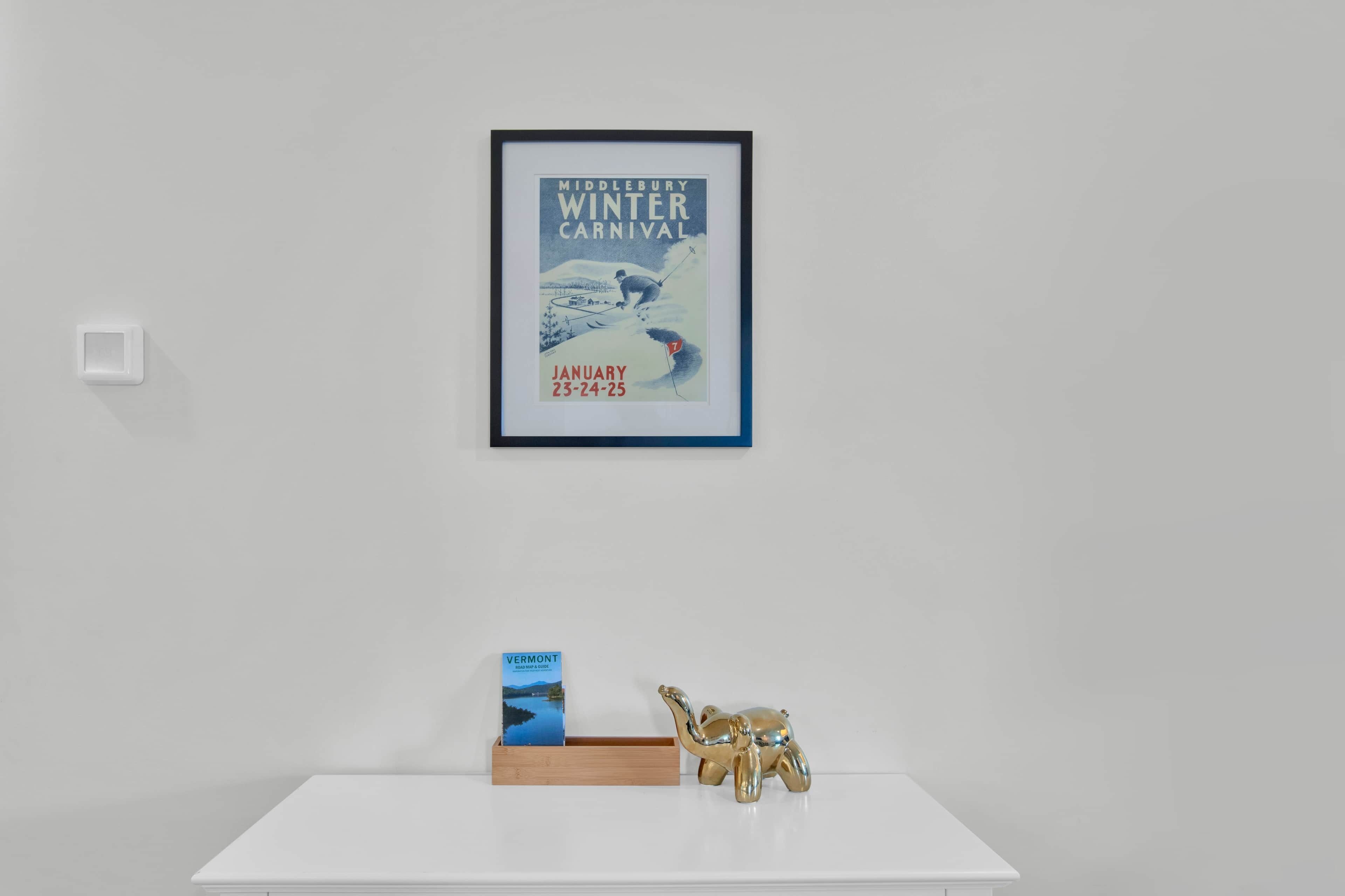 Een lijst met een affiche van een wintercarnaval, een kaart en gids van Vermont, en een gouden olifantbeeldje op een witte plank.