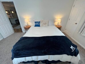 Ein Bett mit einer dunkelblauen Decke, einem Kopfkissen mit dem Schriftzug 'SKI' und zwei Nachttischlampen.