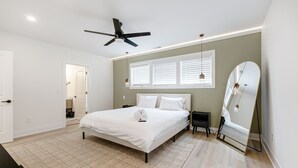 Une chambre à coucher avec un lit, un ventilateur de plafond, un grand miroir et une fenêtre avec des stores.