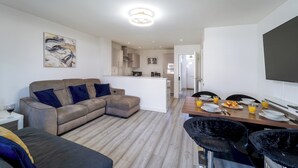 Een moderne woonkamer met een bank, een eettafel met stoelen, een keukenhoek en een flatscreen televisie aan de wand.
